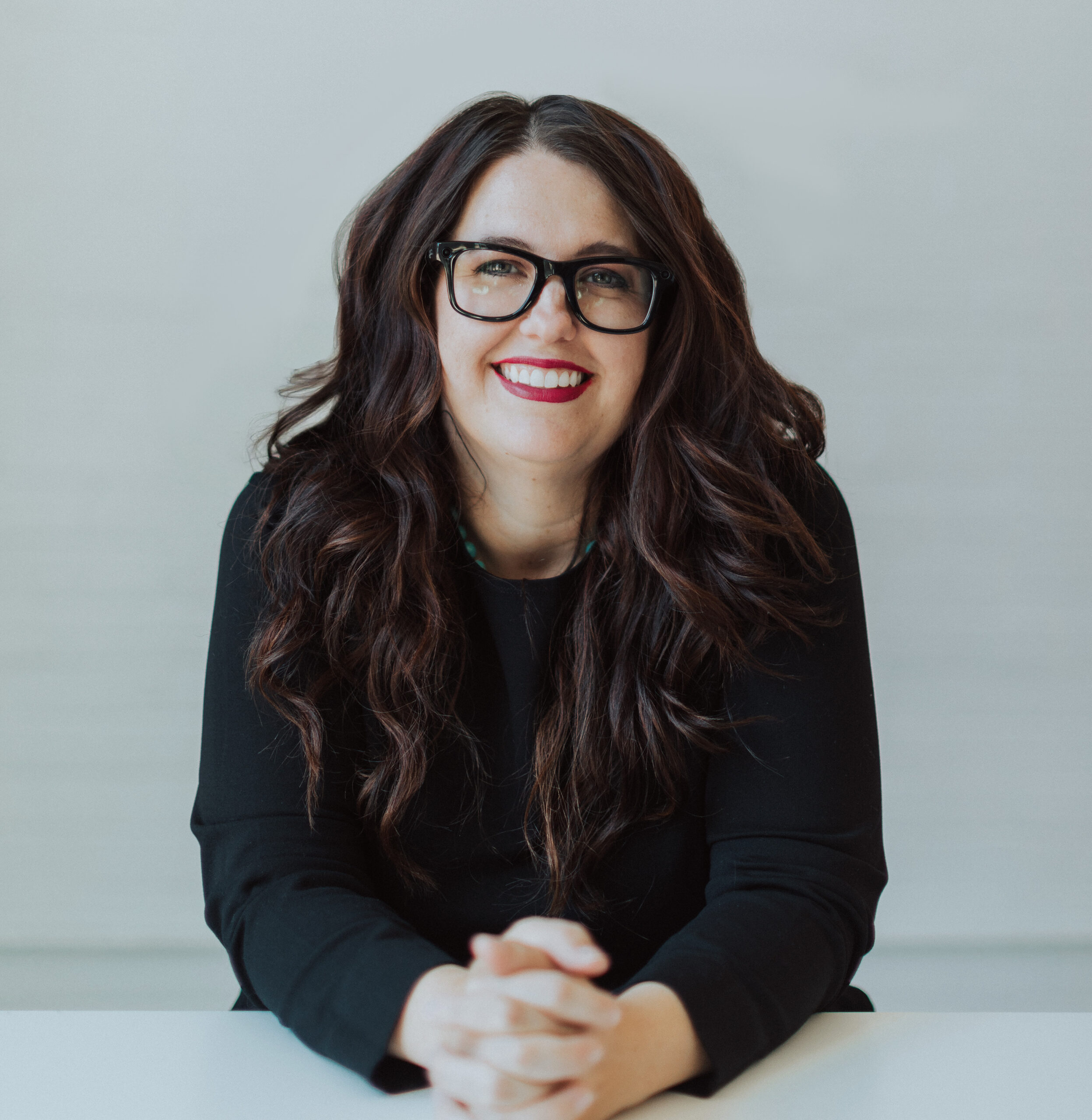 A woman with glasses smiles at the camera while folding her hands together in front of her and leaning forward on a desk.